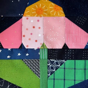 May include: Close-up of a colorful quilt block with various fabric patterns. The design features pink, yellow, green, and blue fabrics with star, sun, and geometric prints. The quilt block is a patchwork of different shapes and textures.