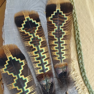 May include: Three turkey feathers with intricate geometric designs painted in gold, green, and black. The feathers are arranged vertically on a wooden surface.