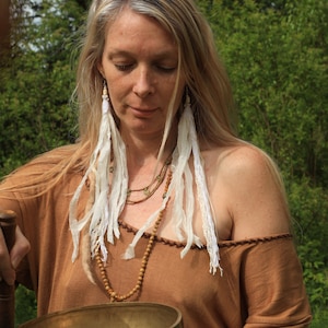 May include: A woman with long blonde hair wearing a brown off-the-shoulder top and a long beaded necklace. She is holding a large, round, brass singing bowl.