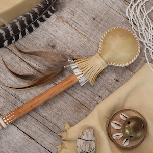 May include: A wooden rattle with a leather head and a fringe of leather tassels. The rattle is decorated with white dots and is lying on a wooden surface with other Native American crafts.