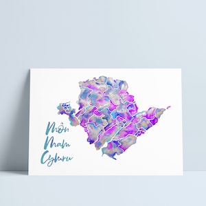 Puede incluir: Una impresión en acuarela de un mapa en tonos morados, azules y plateados, con el texto "Môn Nham Cyhru" en escritura turquesa. El mapa está sobre un fondo blanco.