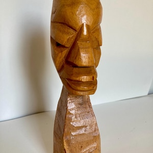 May include: A hand-carved wooden sculpture of a human head and neck. The piece features a light brown finish with visible chisel marks and a smooth, polished surface on the facial features, showcasing the natural grain and texture of the wood.