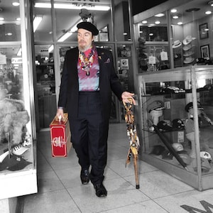 May include: A man wearing a black suit jacket, a patterned shirt, and a black fedora hat is standing in a store. He is holding a red suitcase and a walking stick. The store has a sign that says "Meyer The Hatter".