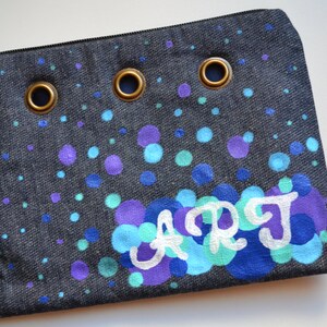 May include: A dark blue denim pouch with a white painted design of the word "ART" surrounded by blue, purple, and teal circles. The pouch has three brass grommets.