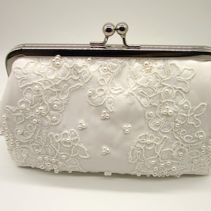 May include: A white satin clutch bag featuring intricate white lace floral appliqués and scattered white pearl beads. The bag has a silver-toned metal frame with a kiss-lock closure at the top, providing a secure opening for the main compartment.
