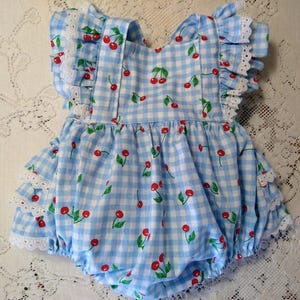 Op de afbeelding: Een babyromper in een blauw en wit geruit patroon, versierd met rode kersenprints. De romper heeft gefranjerde mouwen en afwerking, met witte kanten accenten. Het kledingstuk is ontworpen met een ballonvorm.