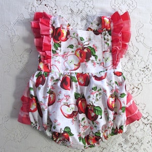 Pode incluir: Um body de bebé branco com estampa de maçãs vermelhas e detalhes florais rosa. O body apresenta mangas com babados e aberturas para as pernas. O tecido é estampado com maçãs vermelhas, folhas verdes e flores rosas.