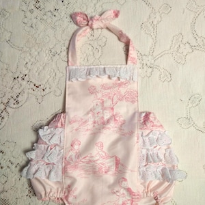 Puede incluir: Un pelele rosa para bebé con ribete de encaje blanco y un estampado toile con una casa, árboles y personas en un barco. El pelele tiene un dobladillo con volantes y un lazo en el cuello.