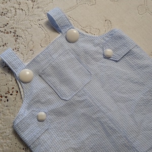 Puede incluir: Pantalones de peto para bebé a rayas azules y blancas con botones de plástico blancos.