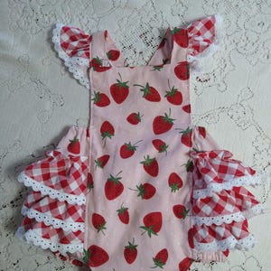May include: A baby romper with a strawberry print on a light pink background. The romper has red and white gingham ruffle sleeves and white lace trim. The straps cross in the back.