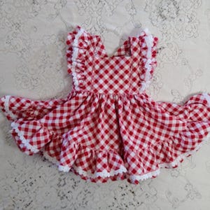 Puede incluir: Un vestido de cuadros rojos y blancos con mangas y dobladillo con volantes. El vestido tiene un escote cuadrado y ribete de encaje blanco. La tela es un patrón de cuadros clásico.