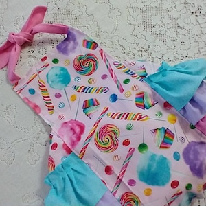 May include: A pink and white baby onesie with a candy theme. The onesie has a pink bow tie at the top and a ruffle trim around the bottom. The fabric features a pattern of lollipops, candy canes, and cotton candy.