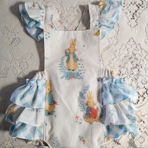 May include: White baby romper with a Peter Rabbit print. The romper features a square neckline, ruffled sleeves, and ruffled trim in a blue and white gingham pattern. The fabric is decorated with illustrations of Peter Rabbit and woodland elements.