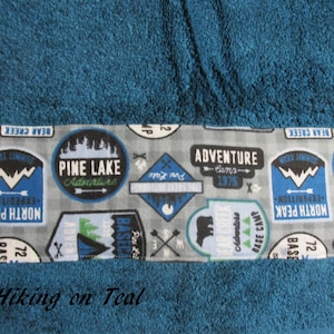 May include: Teal towel with a gray border featuring a repeating pattern of blue and white camping-themed patches. The patches have text that reads "Pine Lake Adventure", "Adventure Camps 1975", "North Peak Expedition", and "Base Camp 72". The text "Hiking on Teal" is printed below the border.