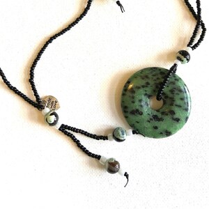 May include: A black beaded necklace with a green and black stone pendant and matching earrings. The pendant is a round disc with a hole in the center. The necklace has a small silver tag that says "Made with Love".