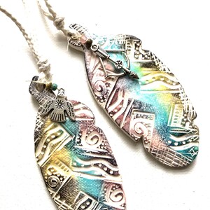 May include: Two colorful ceramic feather pendants with white cord. One pendant features a silver dove charm and the other features a silver arrow charm.