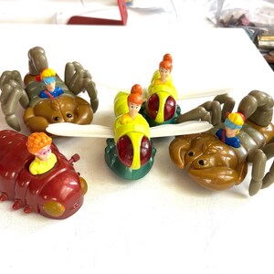 May include: A collection of colorful plastic insect toys, including a brown scorpion, a red caterpillar, and a green and yellow fly. Each toy features a small figure in a vehicle. The toys are likely from a children's toy set.