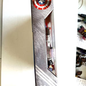 Op de afbeelding: Een kartonnen doos met een zilveren metallic achtergrond en een rood, wit en blauw Amerikaans vlagontwerp. De doos heeft een witte ster en twee witte strepen. De doos is voor een Captain America-actiefiguur.