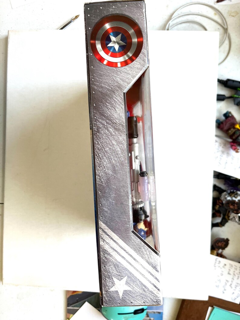 Op de afbeelding: Een kartonnen doos met een zilveren metallic achtergrond en een rood, wit en blauw Amerikaans vlagontwerp. De doos heeft een witte ster en twee witte strepen. De doos is voor een Captain America-actiefiguur.