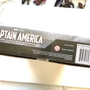 Op de afbeelding: Een kartonnen doos voor een Captain America speelgoed. De doos is zwart en grijs met een afbeelding van Captain America op de voorkant. De tekst "MARVEL STUDIOS" en "CAPTAIN AMERICA THE FIRST AVENGER" staat op de doos gedrukt. De doos heeft ook een waarschuwingslabel met de tekst "Bevat kleine onderdelen." en een barcode.
