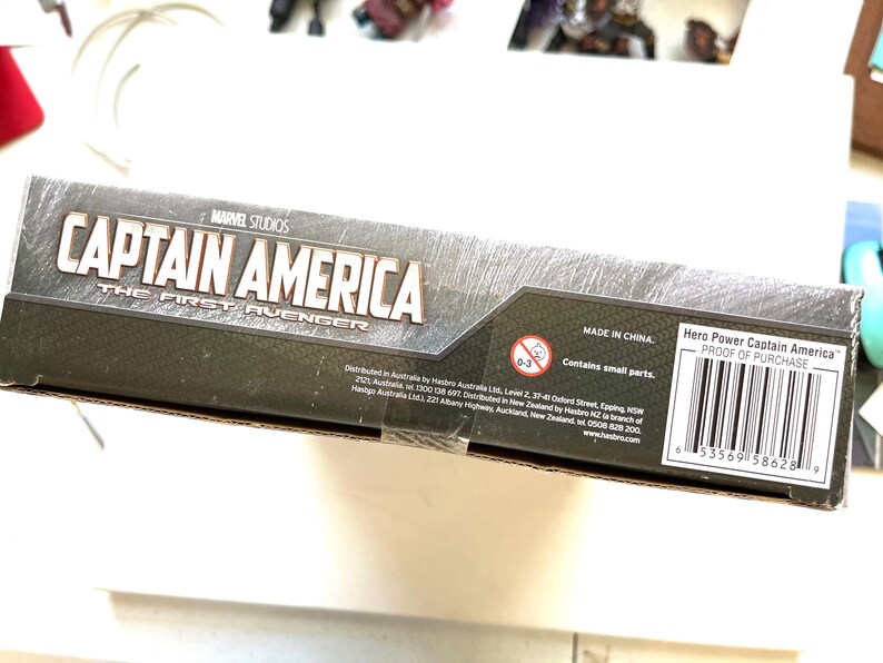 Op de afbeelding: Een kartonnen doos voor een Captain America speelgoed. De doos is zwart en grijs met een afbeelding van Captain America op de voorkant. De tekst "MARVEL STUDIOS" en "CAPTAIN AMERICA THE FIRST AVENGER" staat op de doos gedrukt. De doos heeft ook een waarschuwingslabel met de tekst "Bevat kleine onderdelen." en een barcode.