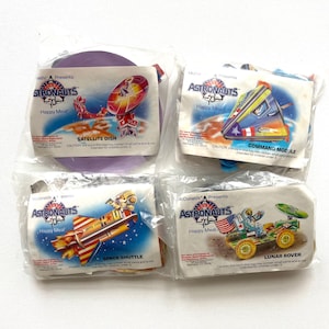 May include: Four sealed McDonald's Happy Meal toys from the Astronauts series. The toys include a purple satellite dish, a command module, a space shuttle, and a lunar rover. Each package features the Astronauts logo and toy name.