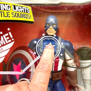 Op de afbeelding: Een plastic action figure van Captain America in zijn iconische rood-wit-blauwe kostuum. De figuur heeft een lichtgevend schild en gevechtsgeluiden. De figuur is verpakt in een doorzichtige plastic doos met een rode en witte achtergrond.