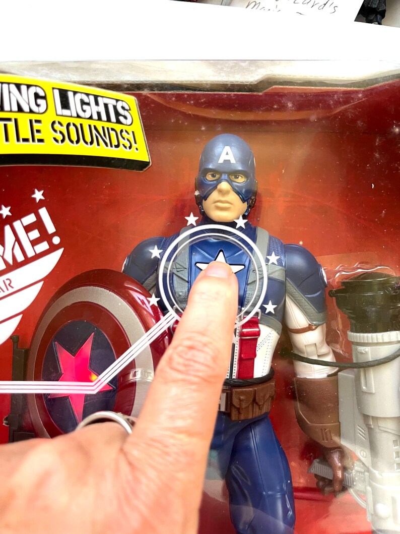 Op de afbeelding: Een plastic action figure van Captain America in zijn iconische rood-wit-blauwe kostuum. De figuur heeft een lichtgevend schild en gevechtsgeluiden. De figuur is verpakt in een doorzichtige plastic doos met een rode en witte achtergrond.