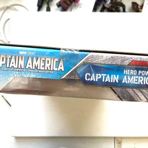 Op de afbeelding: Een blauwe en rode doos met de tekst "Captain America" en "The First Avenger" op de voorkant. De doos heeft ook de tekst "Hero Power Captain America" op de voorkant.