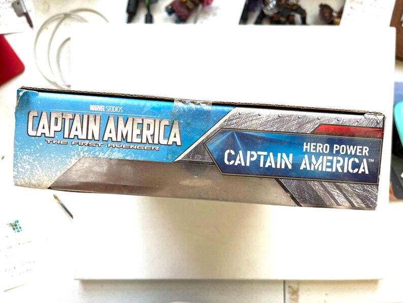 Op de afbeelding: Een blauwe en rode doos met de tekst "Captain America" en "The First Avenger" op de voorkant. De doos heeft ook de tekst "Hero Power Captain America" op de voorkant.