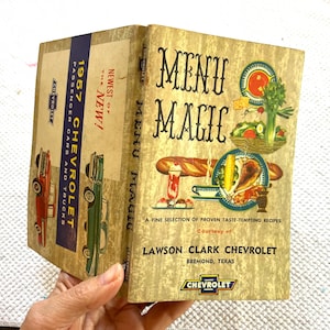 May include: Vintage cookbook titled "Menu Magic" with illustrations of food. The cover features the text "Menu Magic" and "A Fine Selection of Proven Taste-Tempting Recipes." The spine reads "1957 Chevrolet Passenger Cars and Trucks."