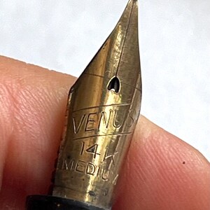 Może przedstawiać: Zbliżenie złotego pi&oacute;ra wiecznego. Na stal&oacute;wce wygrawerowano słowa "VENUS", "14K" i "MEDIUM". Pi&oacute;ro ma czarny korpus. Pi&oacute;ro jest trzymane przez osobę.