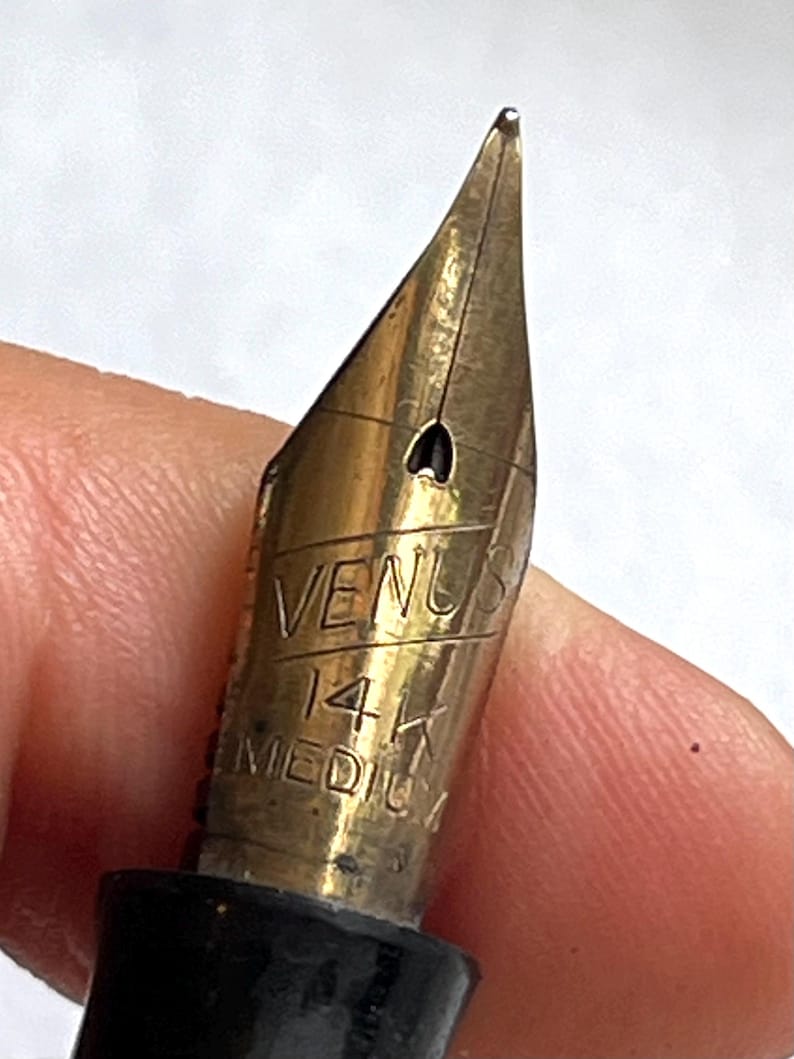 Może przedstawiać: Zbliżenie złotego pi&oacute;ra wiecznego. Na stal&oacute;wce wygrawerowano słowa "VENUS", "14K" i "MEDIUM". Pi&oacute;ro ma czarny korpus. Pi&oacute;ro jest trzymane przez osobę.