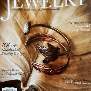 Może przedstawiać: Okładka magazynu Belle Armoire Jewelry przedstawiająca zbliżenie ręcznie robionych projekt&oacute;w biżuterii wykonanych z lampworkowych szklanych koralik&oacute;w, polimerowej gliny i drutu. Na okładce widnieje tekst "Belle Armoire Jewelry" i "100+ ręcznie robionych projekt&oacute;w biżuterii".