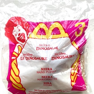 May include: A plastic bag with a pink and yellow design featuring the McDonald's logo and the words "Walt Disney Dinosaur" and "Le Dinosaure". The bag contains a brown hand puppet with the text "Neera Hand Puppet", "Neera Marionnette", and "Nira Titere" printed on it.