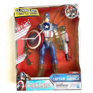 Op de afbeelding: Captain America actionfiguur met gloeiende lichten en gevechtsgeluiden. De figuur houdt een schild en een pistool vast. De verpakking heeft het Avengers Assemble logo en de woorden "Hero Power".