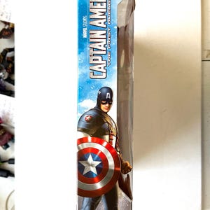 Op de afbeelding: Een Captain America action figure in een doos. De figuur draagt een rood, wit en blauw kostuum met een schild. De doos heeft een blauwe achtergrond met de woorden "Captain America" en "The First Avenger" in wit.