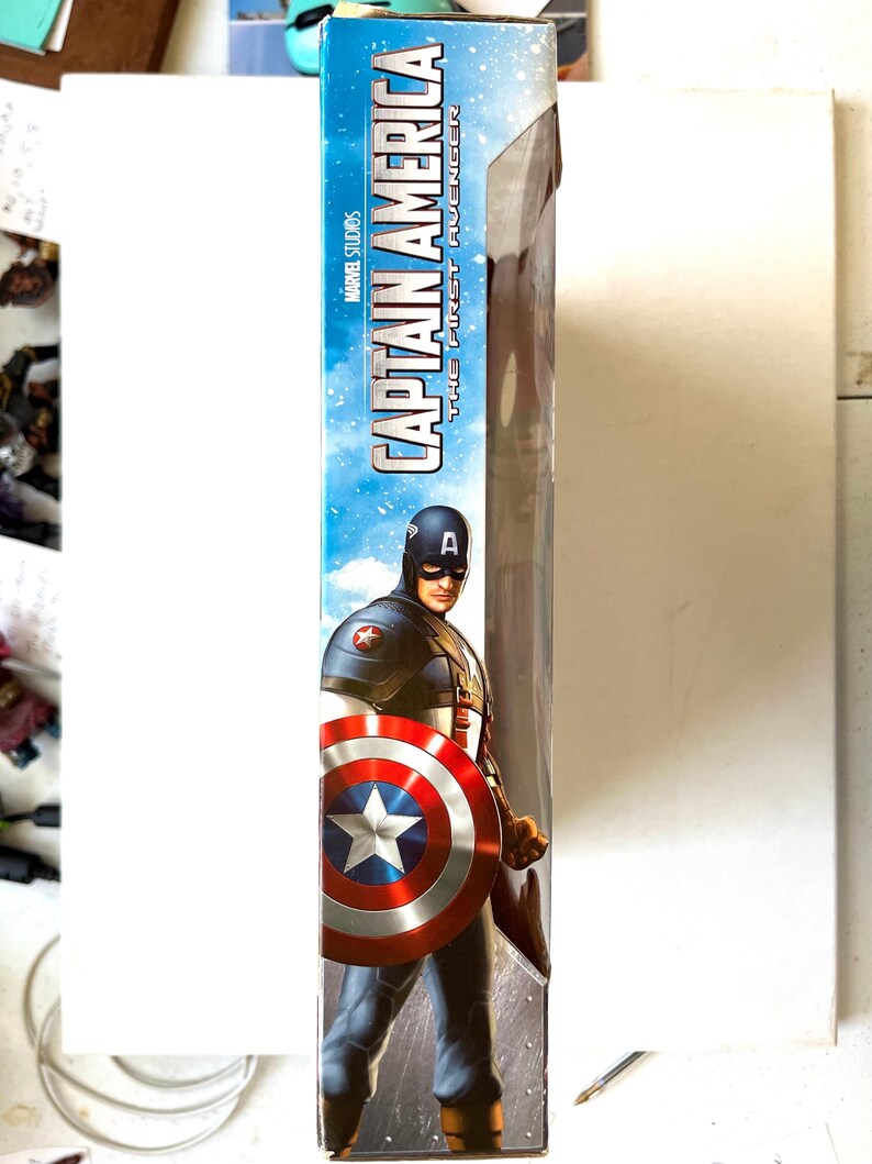 Op de afbeelding: Een Captain America action figure in een doos. De figuur draagt een rood, wit en blauw kostuum met een schild. De doos heeft een blauwe achtergrond met de woorden "Captain America" en "The First Avenger" in wit.