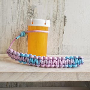 May include: A light blue and pink paracord lanyard with a white pill bottle attached. The lanyard is made of braided cord and has a knot at the top.