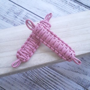 May include: Two pink paracord handles, woven with a tight, intricate pattern. The handles are made of durable, braided cord and have a loop at each end.