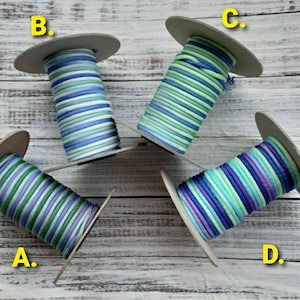 May include: Four spools of colorful cord, each wound on a cardboard spool. The cords display various color combinations, including blue, green, purple, and white. The spools are arranged on a light-colored wooden surface.