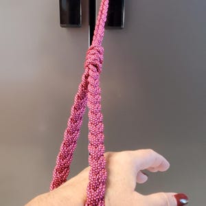 May include: A braided lanyard made of pink and black cord with a repeating speckled pattern. The cord is formed into a loop with a knot at the top, designed to be worn around the wrist. The texture is consistent throughout the braided structure.