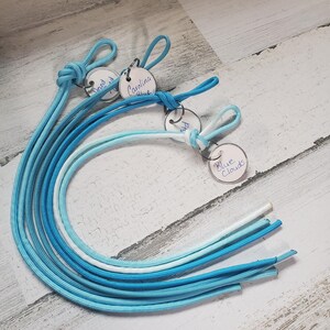 May include: A collection of blue and white cords with knotted loops and small, round tags. The tags are labeled with handwritten text, including "Carolina Blue" and "Blue Clouds". The cords vary in shades of blue.