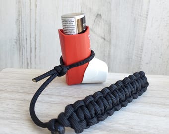 May include: A red and white inhaler with a black paracord lanyard. The inhaler is secured with the lanyard, which is knotted around the device. The lanyard has a woven design.