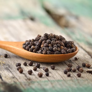 May include: A wooden spoon filled with black peppercorns sits on a rustic wooden surface. Scattered peppercorns are visible around the spoon.