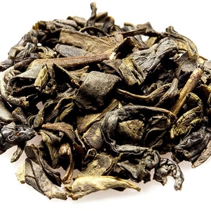 May include: Loose green tea leaves, some with a darker brown hue, are scattered on a white background. The leaves are dried and ready to be brewed.