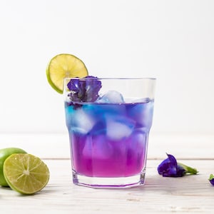 May include: A glass of blue and purple butterfly pea flower tea with ice and a lime wedge garnish. The drink is on a white wooden surface with fresh butterfly pea flowers.