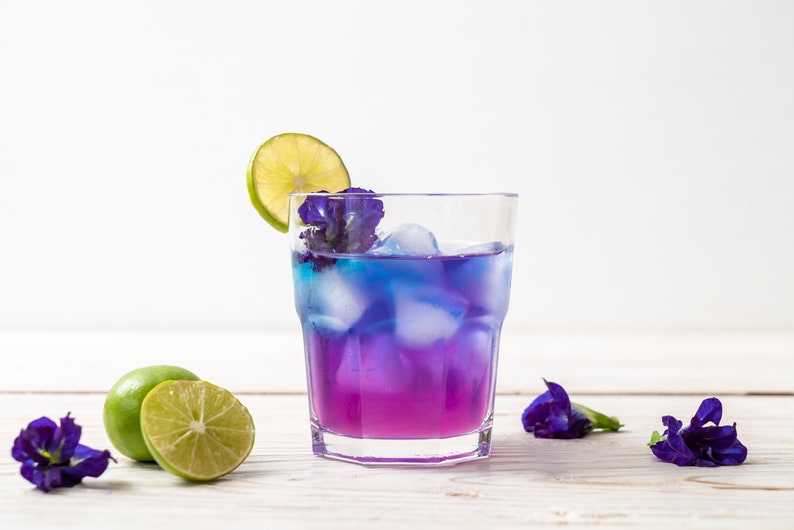 May include: A glass of blue and purple butterfly pea flower tea with ice and a lime wedge garnish. The drink is on a white wooden surface with fresh butterfly pea flowers.