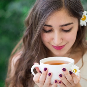 May include: A woman with long brown hair is holding a white teacup with a light brown tea inside. She is wearing a white tank top and has a white daisy in her hair.