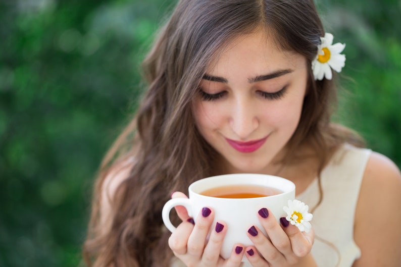 May include: A woman with long brown hair is holding a white teacup with a light brown tea inside. She is wearing a white tank top and has a white daisy in her hair.